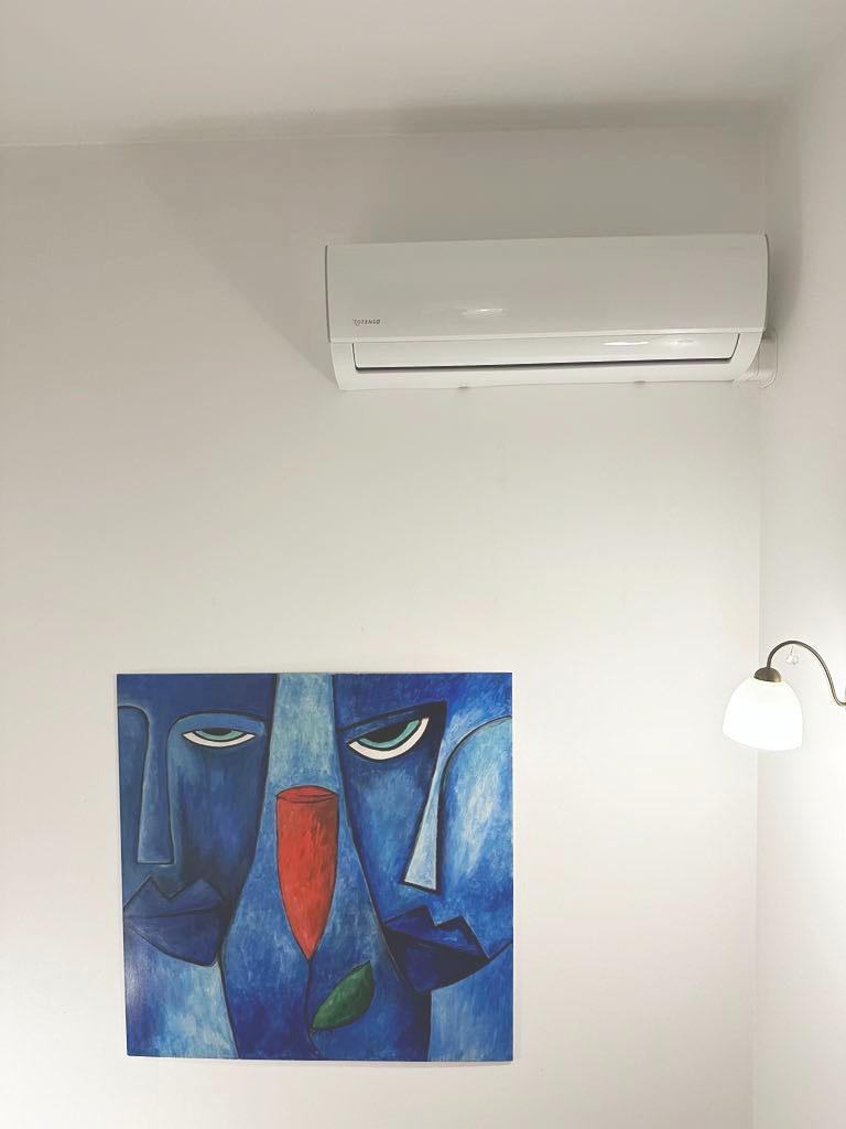 Biały klimatyzator na ścianie nad abstrakcyjnym obrazem w odcieniach błękitu i czerwieni, oświetlony ciepłym światłem lampki.