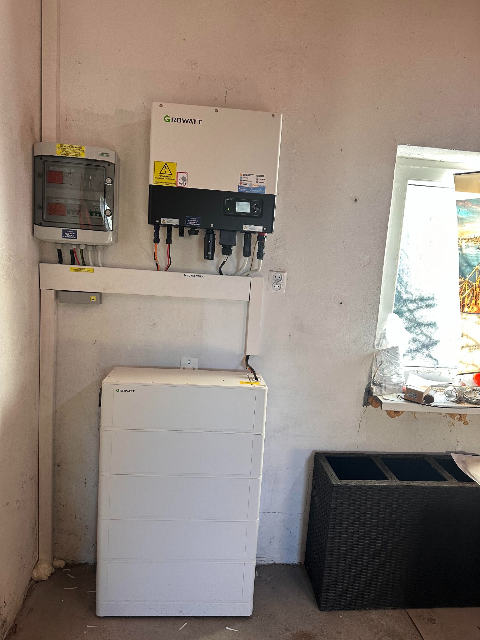 Instalacja fotowoltaiczna w pomieszczeniu: inwerter Growatt, skrzynka z bezpiecznikami i akumulator. Widoczne okablowanie i elementy montażowe.