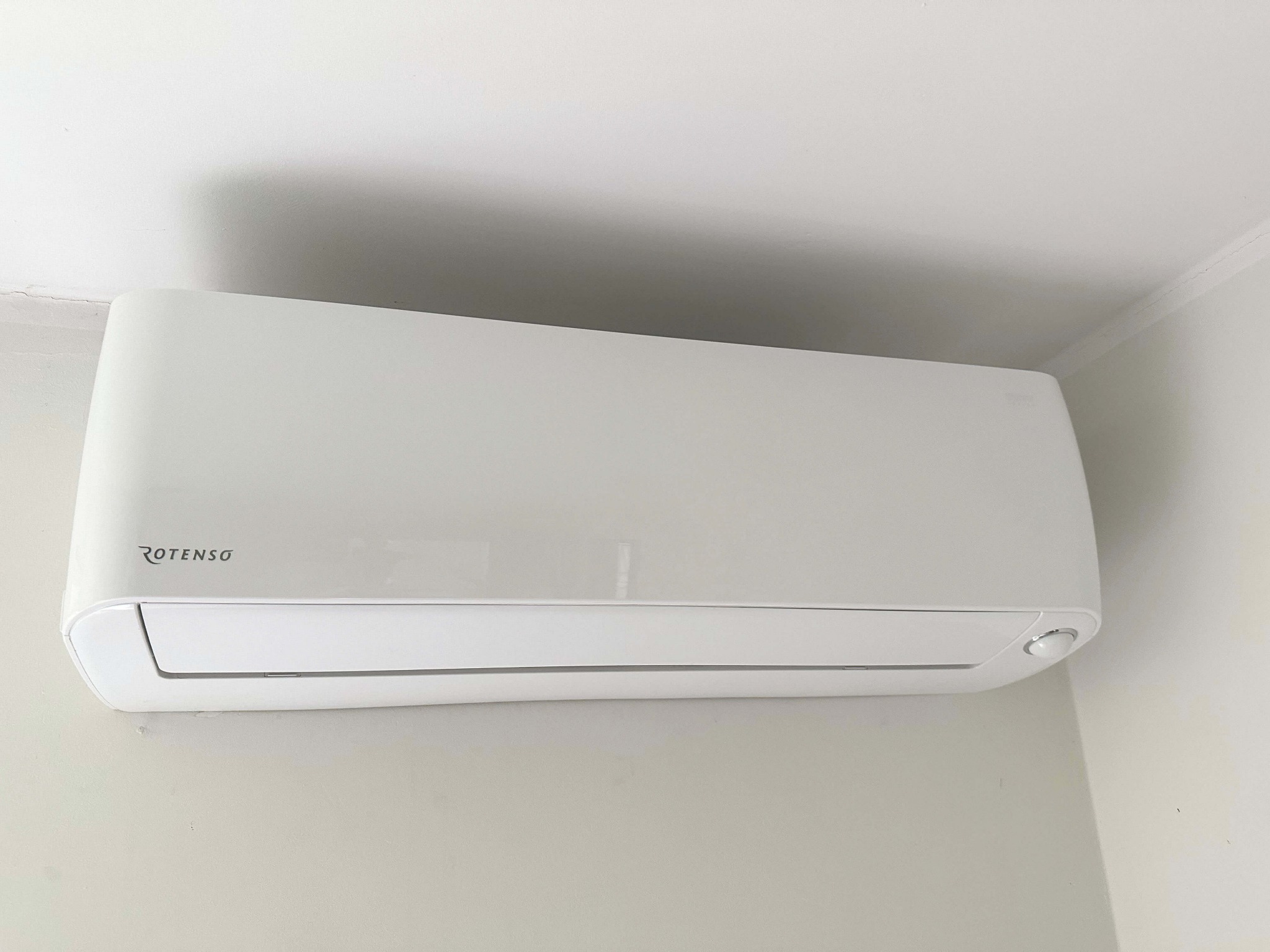 Biała jednostka klimatyzacji Rotenso zamontowana na ścianie w jasnym pomieszczeniu. Widoczny fragment sufitu i ściany, urządzenie w centralnej części kadru.
