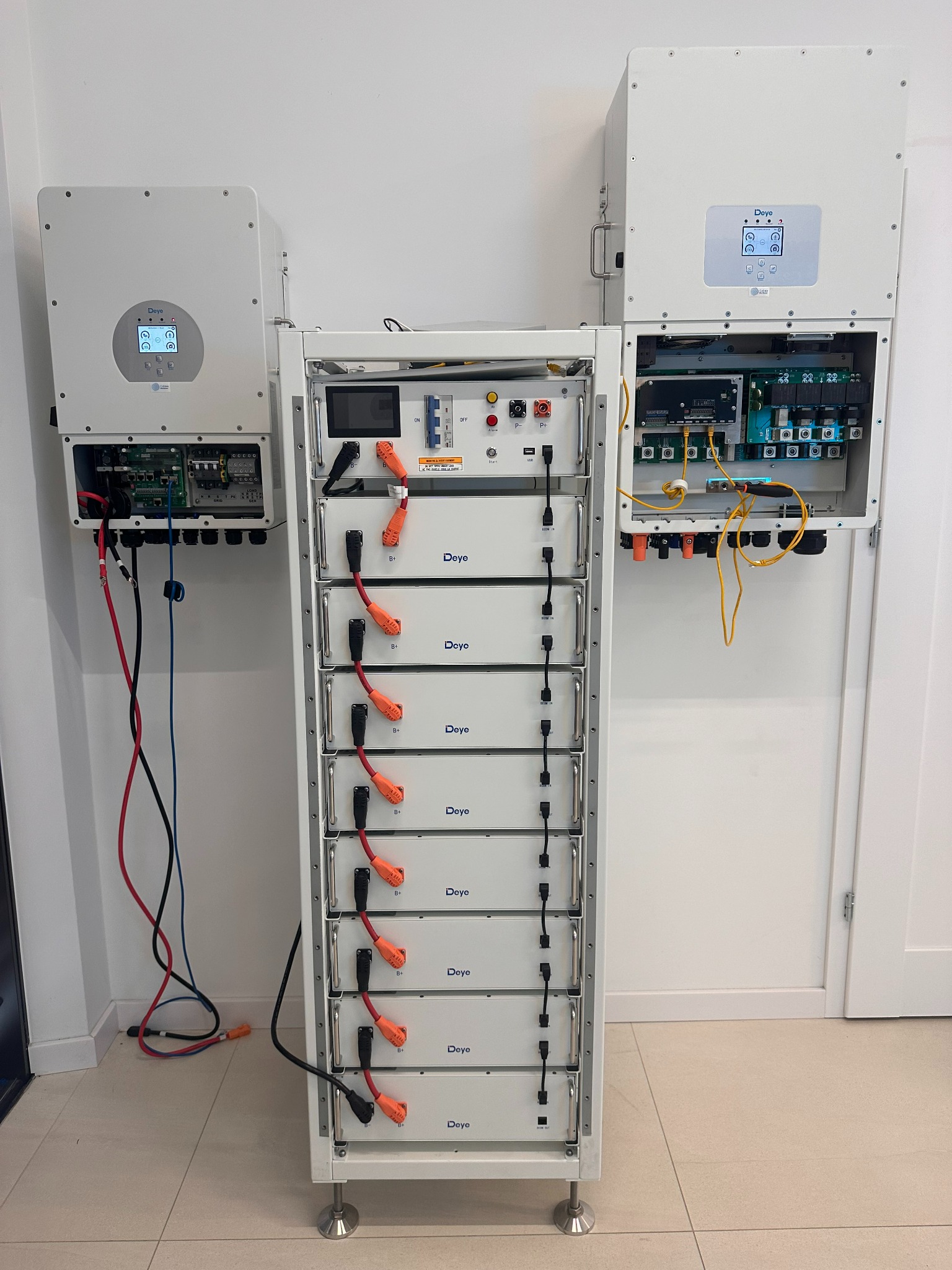 Biała szafa z modułami bateryjnymi Deye, zasilana z dwóch białych inwerterów na ścianie. Widoczne okablowanie i podłączenia. Całość w jasnym, minimalistycznym wnętrzu.