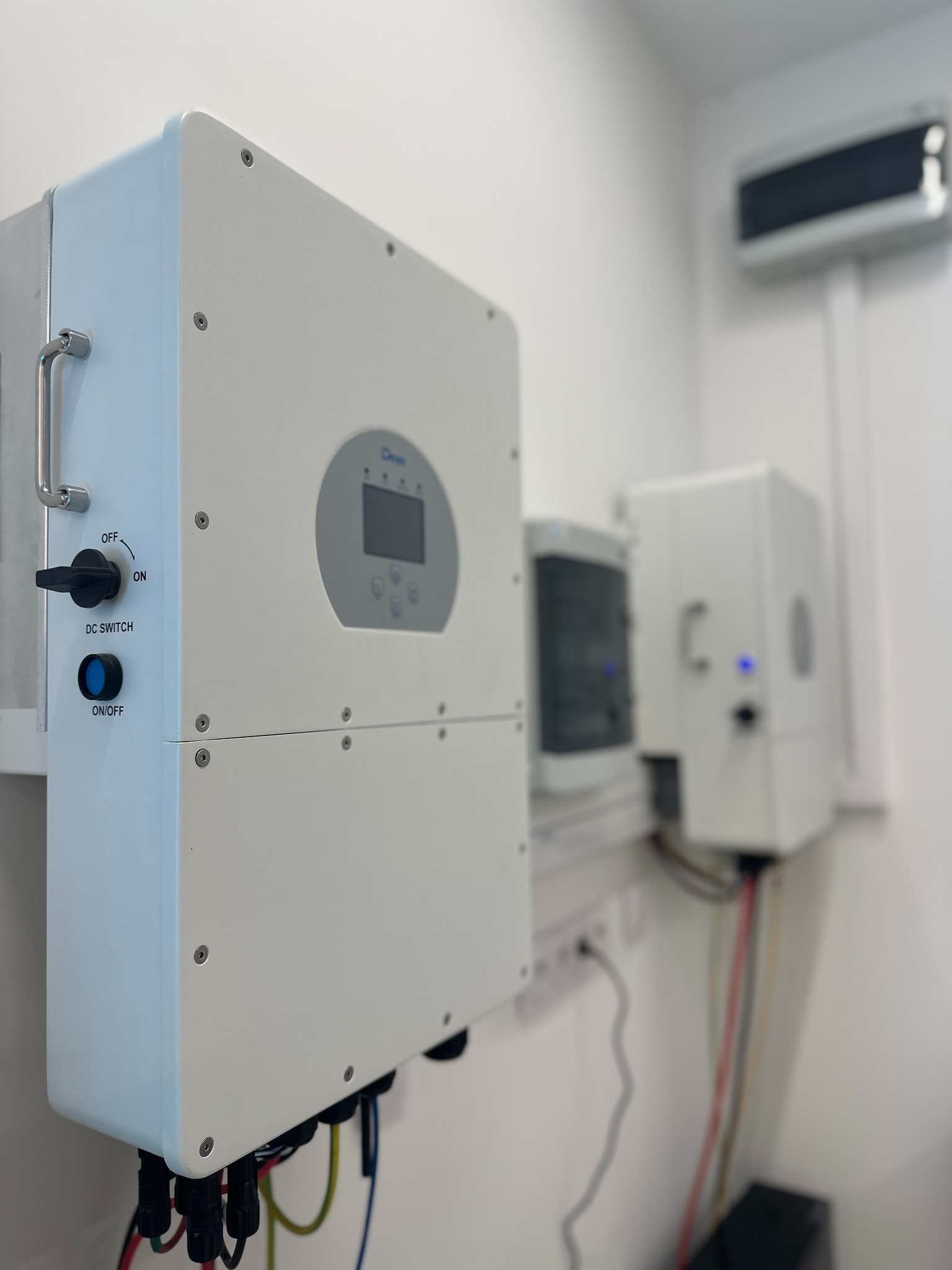 Biały inwerter fotowoltaiczny z przełącznikiem DC i wyświetlaczem, zamontowany na ścianie w pomieszczeniu. Widoczne kable i elementy instalacji.