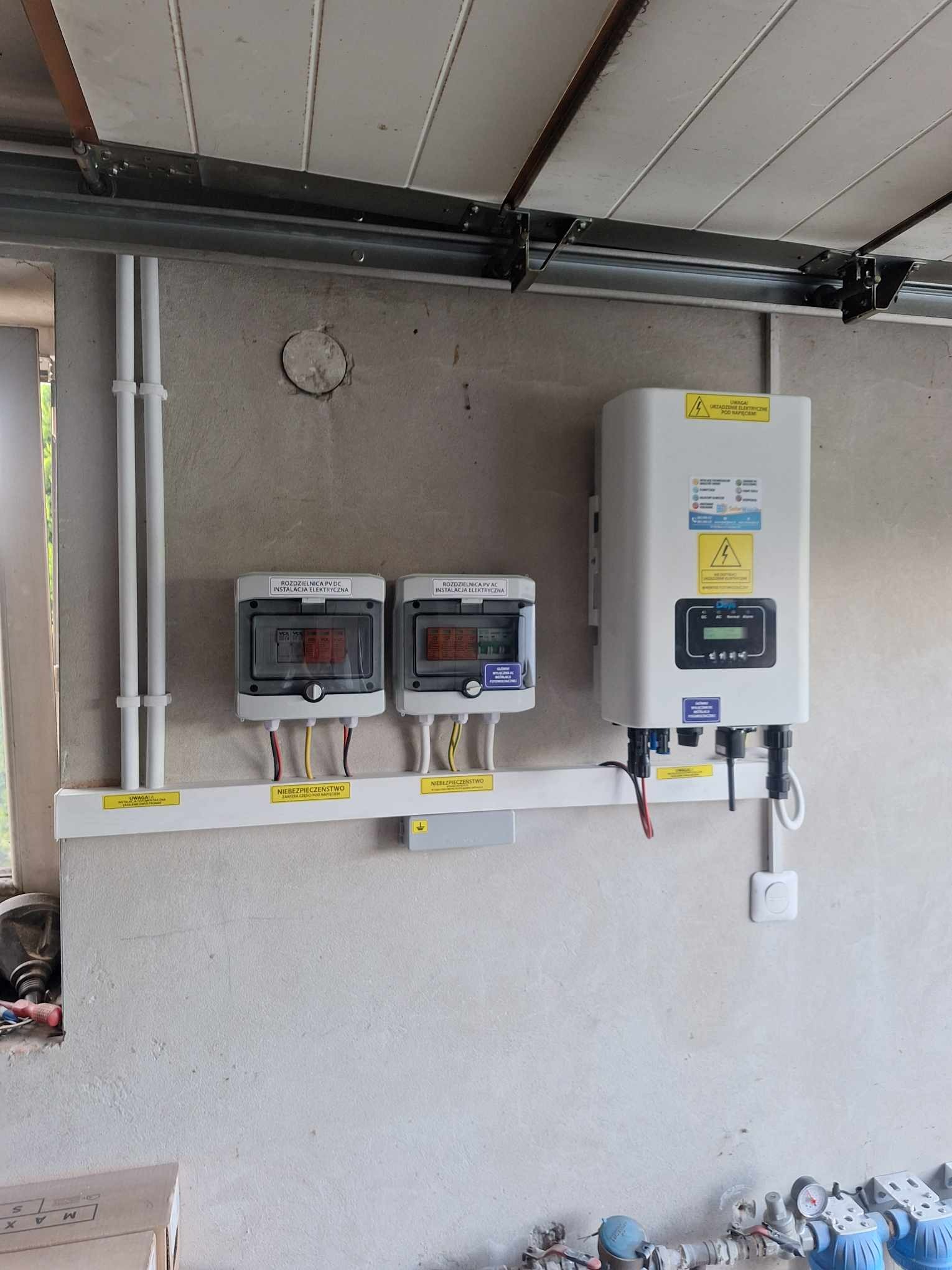 Instalacja fotowoltaiczna w garażu: dwa rozdzielnice PV DC/AC i inwerter na ścianie, pod sufitem brama garażowa. Widoczne przewody i oznaczenia ostrzegawcze.