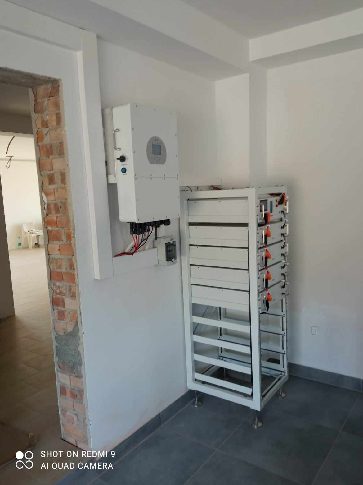Biały inwerter fotowoltaiczny i moduł magazynu energii w surowym wnętrzu. Widoczne kable i instalacja elektryczna. Szare płytki podłogowe i ceglana ściana w tle.