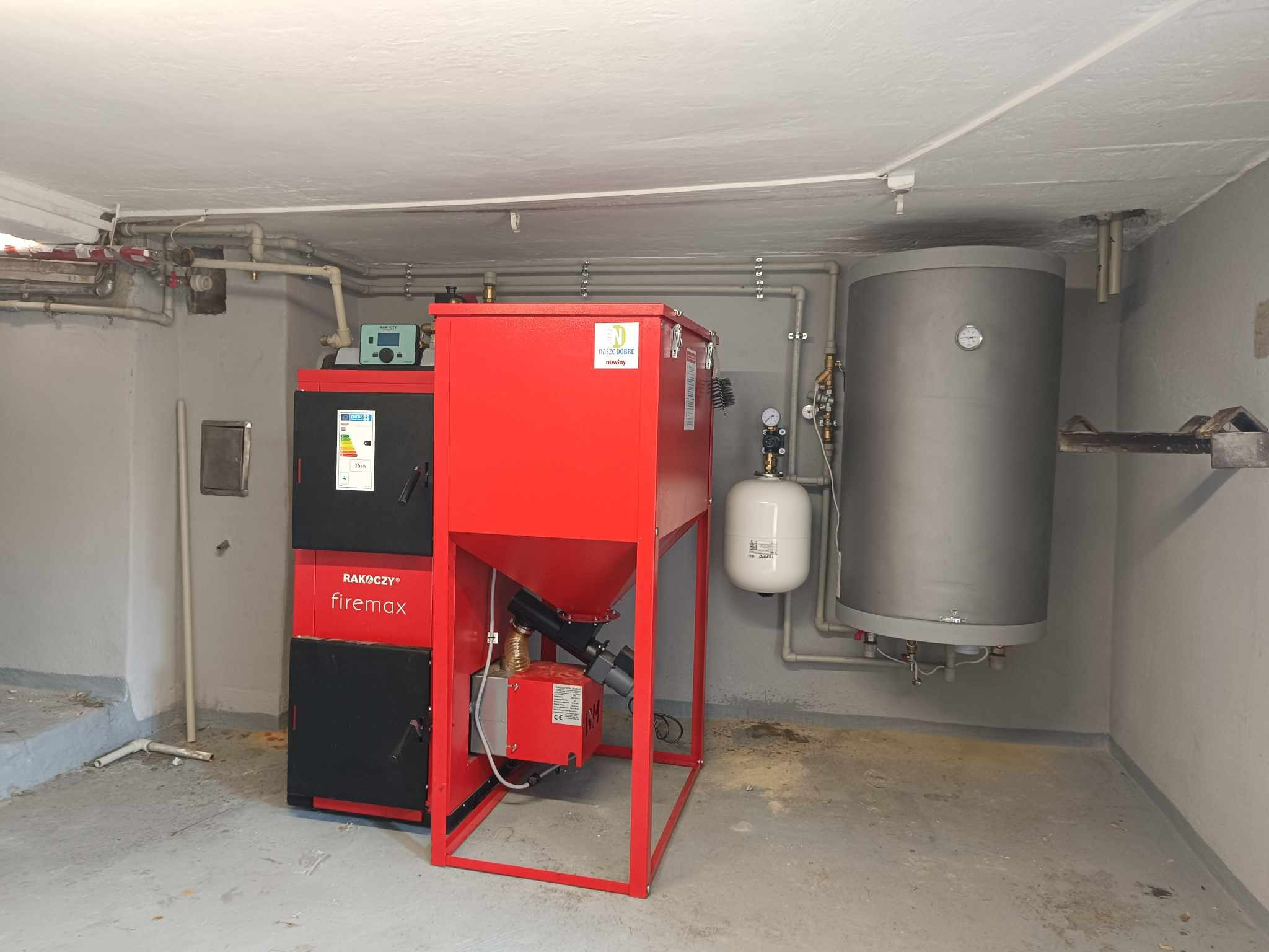 Kompletna instalacja kotła na paliwo stałe Rakoczy Firemax z zasobnikiem i zbiornikiem wyrównawczym w kotłowni, z widocznymi rurami instalacji grzewczej.