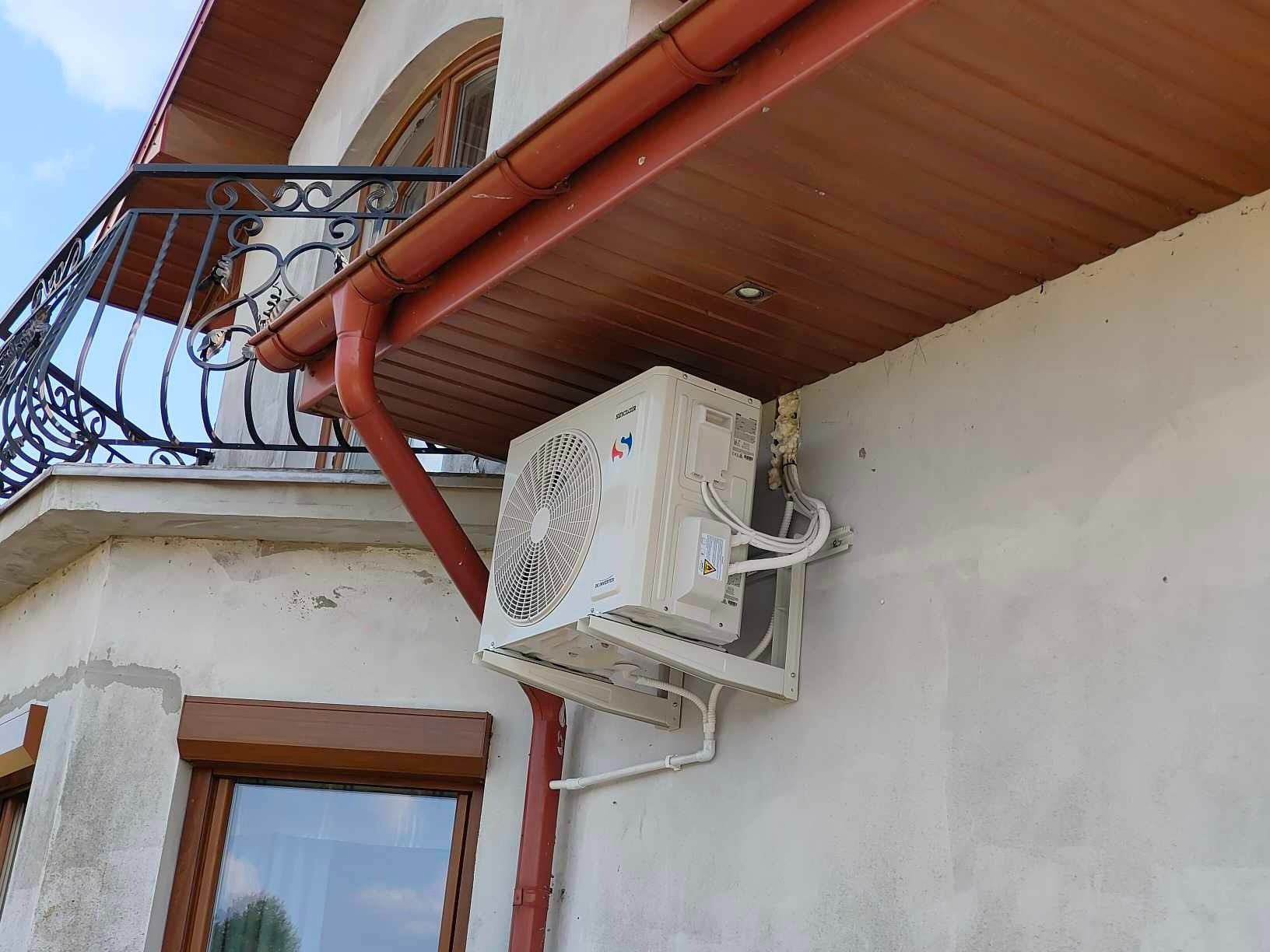 Biały klimatyzator na elewacji domu z brązowymi rynnami i balkonem z kutą balustradą. Widoczne przewody i pianka montażowa. Ujęcie z dołu.