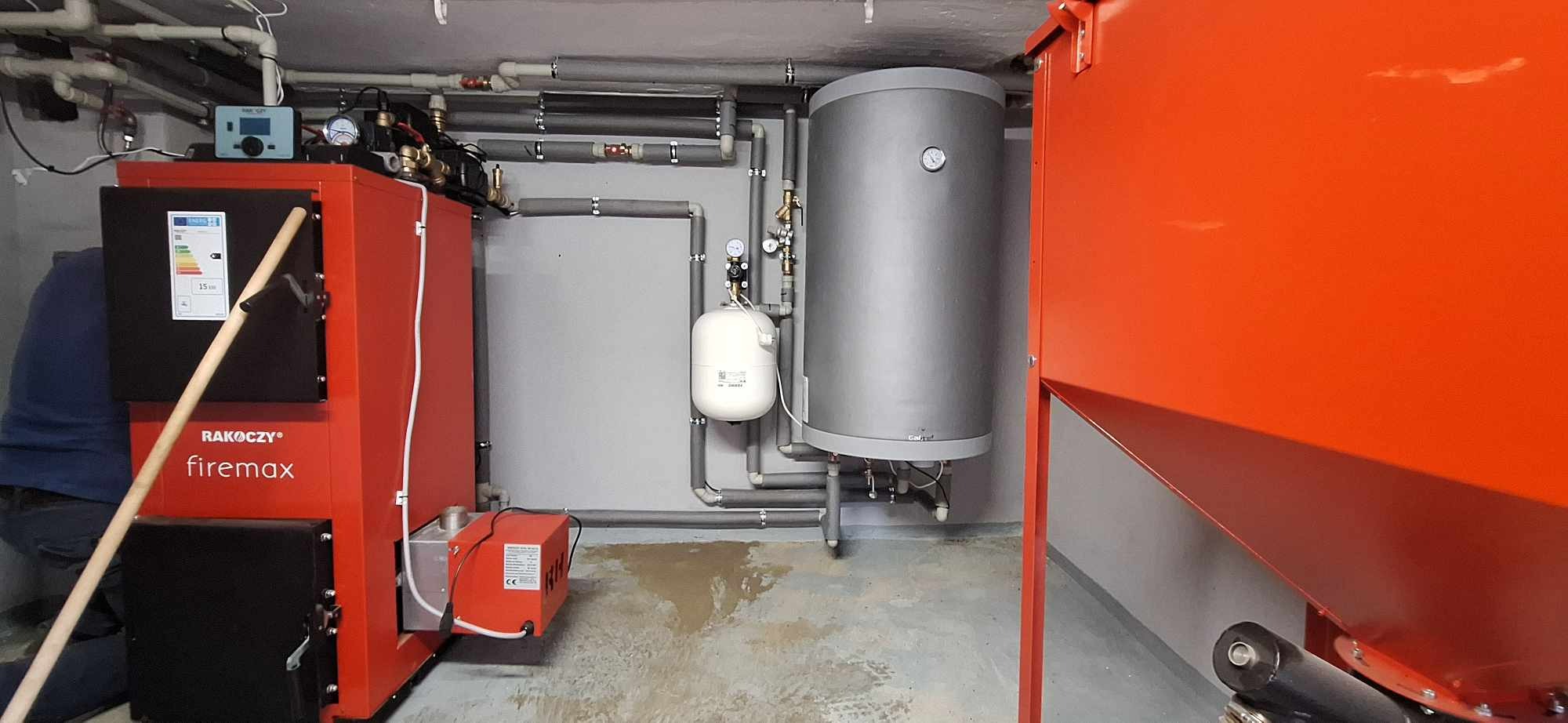Czerwony piec marki Rakoczy Firemax z osprzętem, zbiornik buforowy i instalacja grzewcza z szarych rur w kotłowni, widok na całość instalacji.