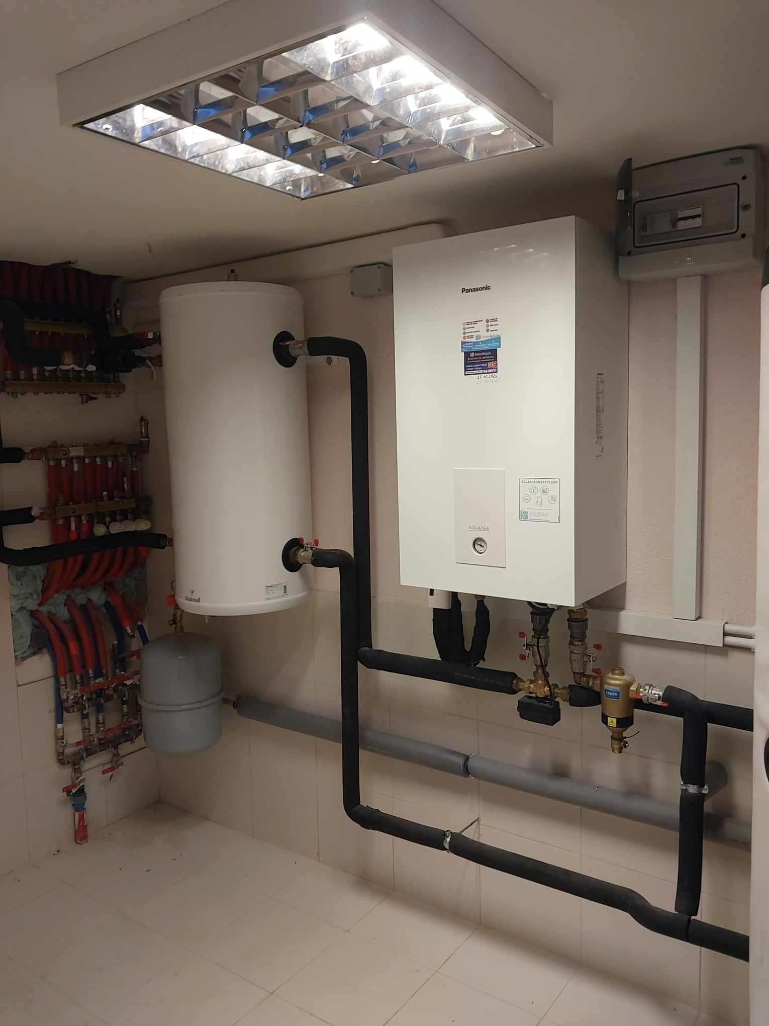 Biały kondensacyjny kocioł gazowy Panasonic Aquarea i zasobnik ciepłej wody, czarne izolowane rury w kotłowni z białymi kaflami, oświetlenie rastrowe.