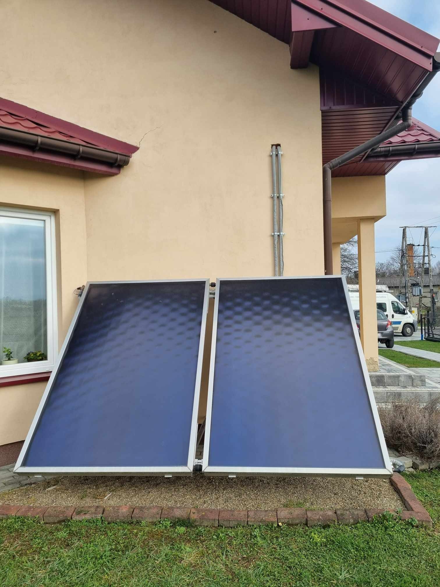 Dwa płaskie kolektory słoneczne zamontowane przy ścianie domu z beżową elewacją, widoczne okno i fragment dachu. Tło stanowi trawnik i fragment ulicy.
