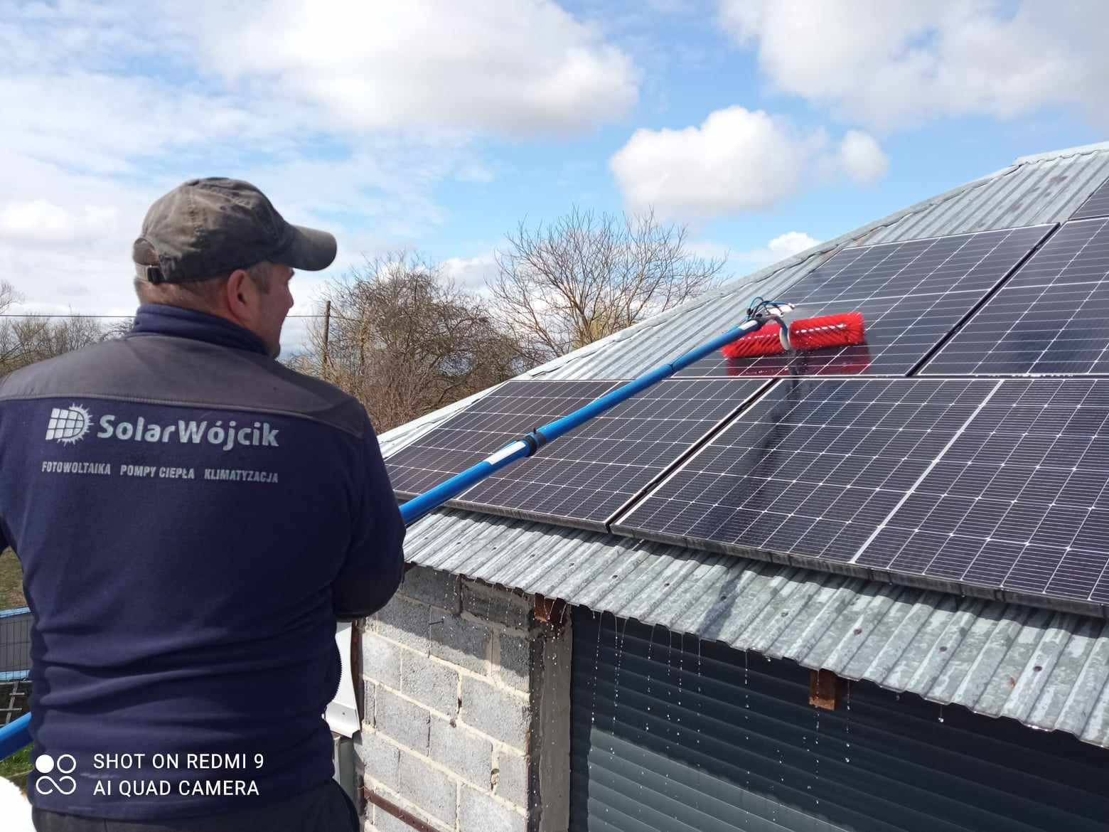 Mężczyzna w uniformie SolarWójcik czyści czerwoną szczotką panele fotowoltaiczne na blaszanym dachu. W tle błękitne niebo i drzewa.