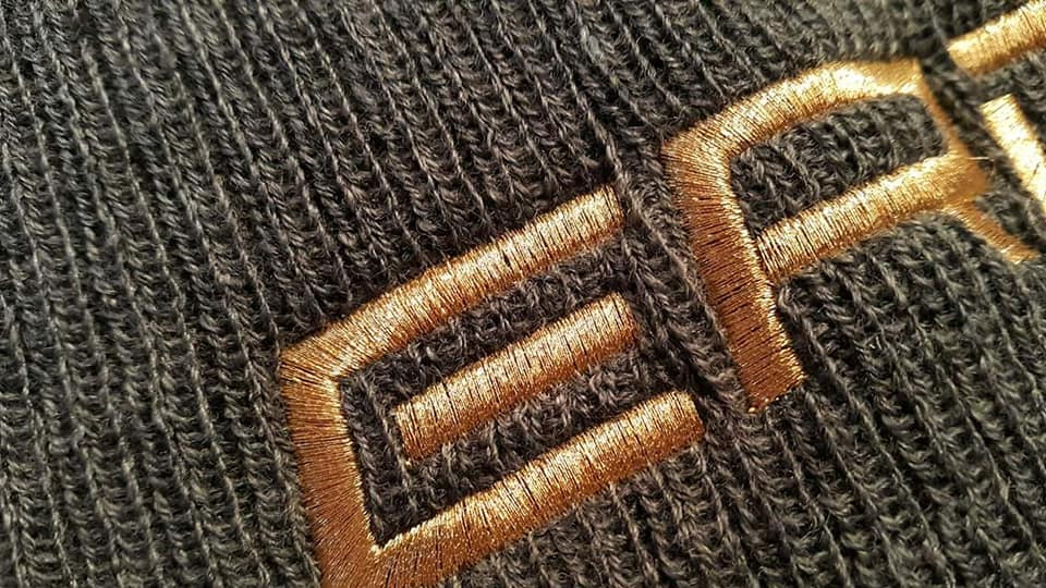 Zbliżenie na szarą dzianinę z wyhaftowanym złotym wzorem geometrycznym.