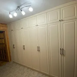 Długa, kremowa zabudowa szafowa z ciemnymi uchwytami, sięgająca sufitu, w pomieszczeniu z drewnianą boazerią i otwartymi drzwiami do kolejnego pokoju.