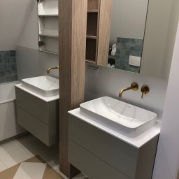 Kuchnie na wymiar | Łukasz Zawodnik - Nowoczesna łazienka z dwoma umywalkami nablatowymi, złotą armaturą i szafkami w odcieniach szarości. Na podłodze wzór w szachownicę. Widoczne lustro i półki.