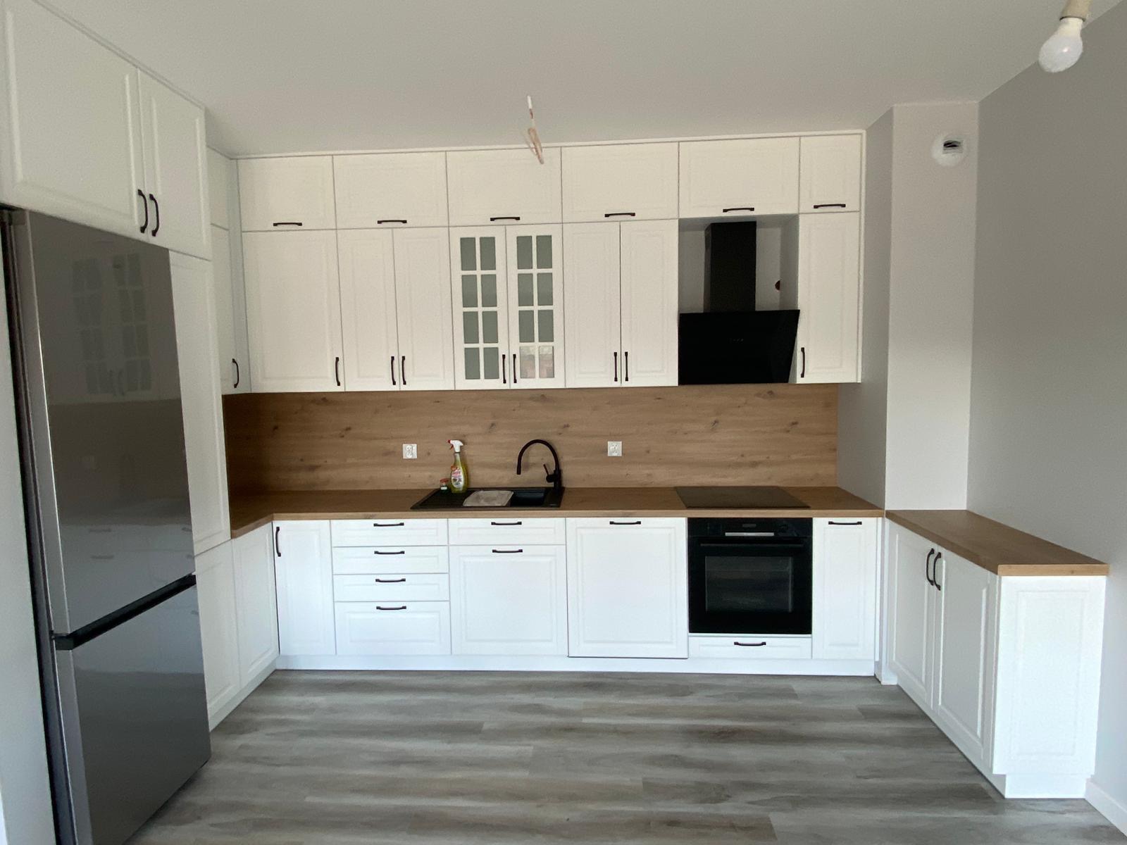 Nowoczesna, jasna kuchnia z białymi szafkami i drewnianym blatem, czarnymi uchwytami i zlewozmywakiem, z widoczną lodówką ze stali nierdzewnej i płytą indukcyjną.