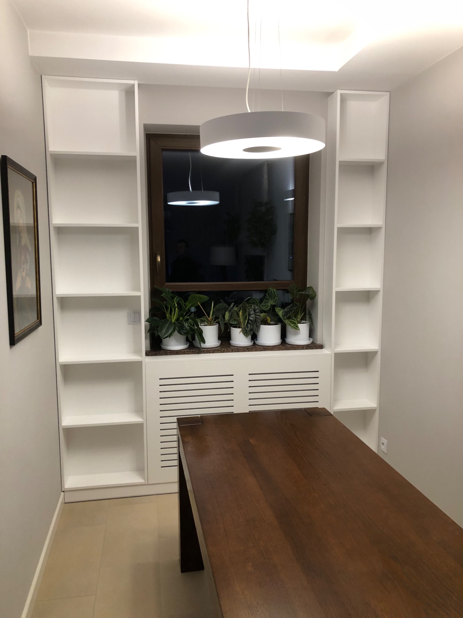 Białe regały wnękowe po bokach okna z roślinami w doniczkach, drewniany blat stołu na pierwszym planie, Kolbuszowa.