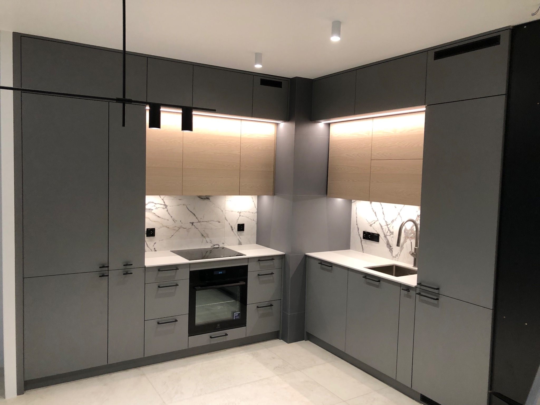 Nowoczesna kuchnia w szarościach z drewnianymi akcentami, oświetleniem LED podszafkowym i marmurowym backsplash'em, widok na zabudowę narożną z piekarnikiem i zlewem.