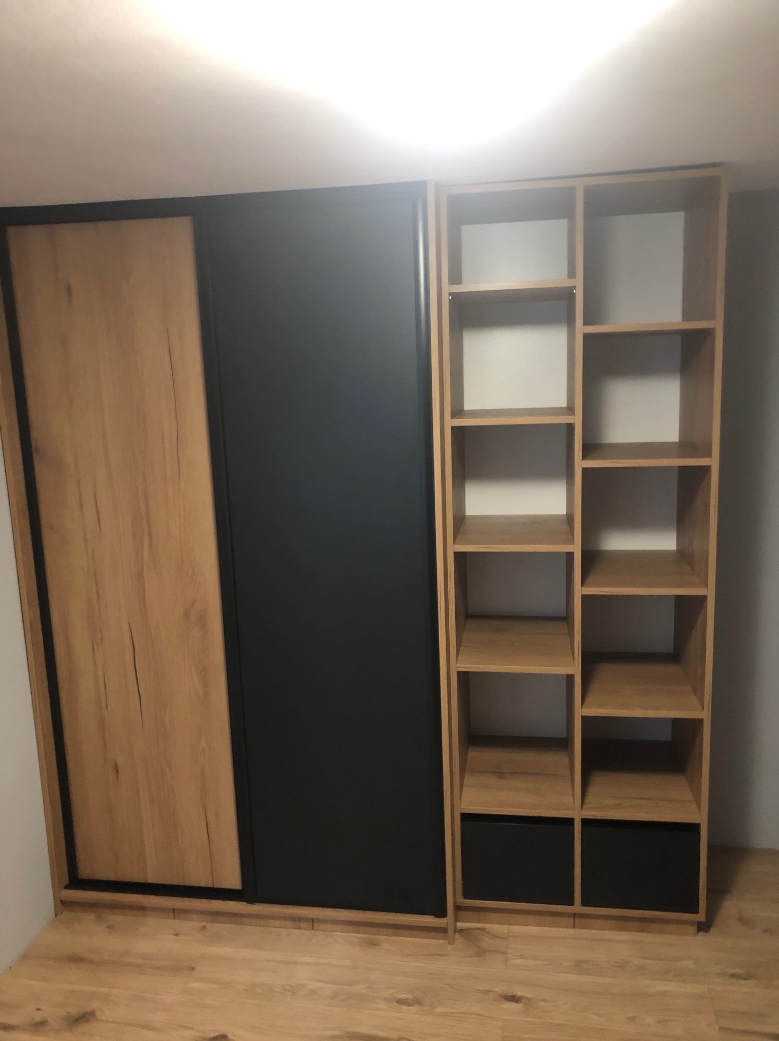 Szafa przesuwna z frontem drewnopodobnym i czarnym, obok regał z półkami i szufladami w kolorach drewna i czerni, na podłodze panele drewnopodobne.