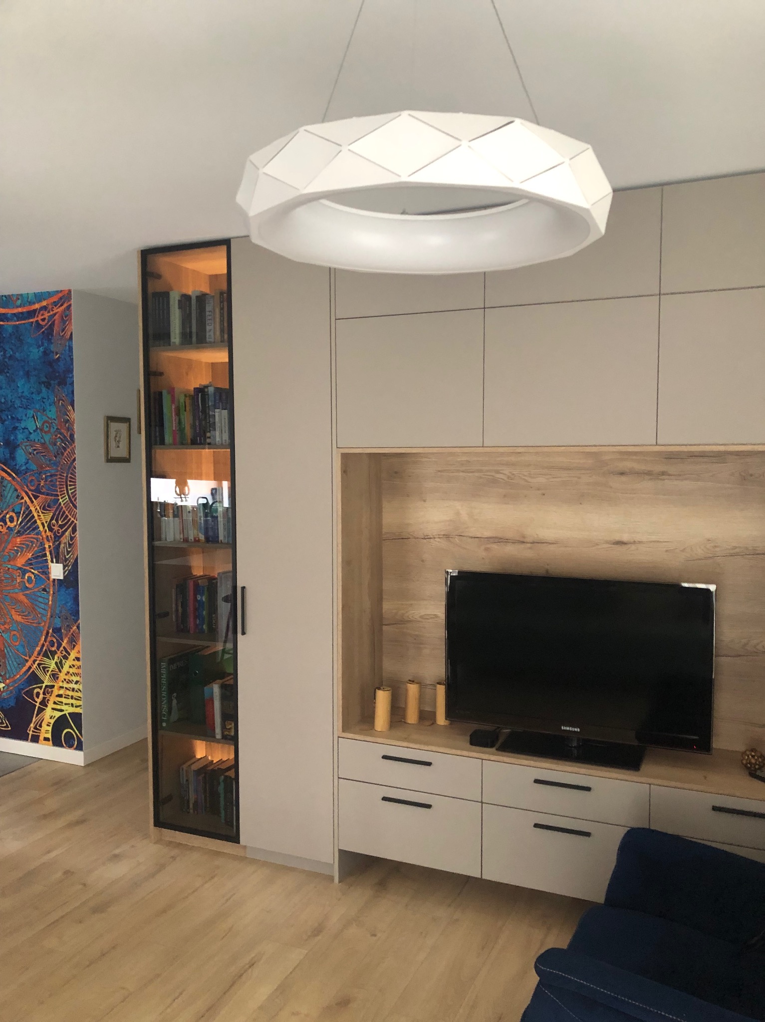 Nowoczesna zabudowa salonu z szafkami w odcieniach bieli i drewna, podświetlana witryna z książkami, okrągła lampa sufitowa i fragment wzorzystej tapety.