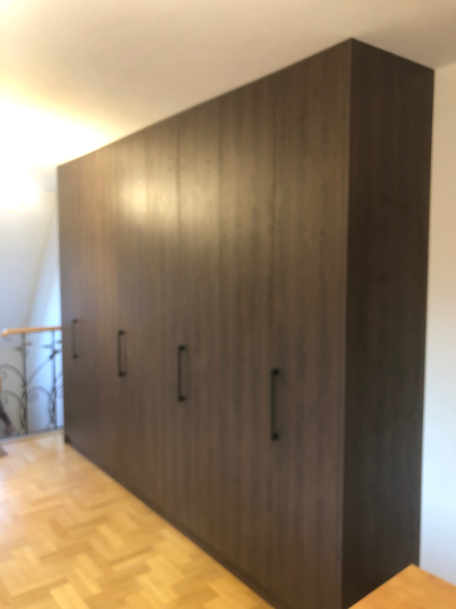 Duża, brązowa szafa wnękowa z sześcioma drzwiami i czarnymi uchwytami, widoczna z boku na tle parkietu i balustrady schodów.