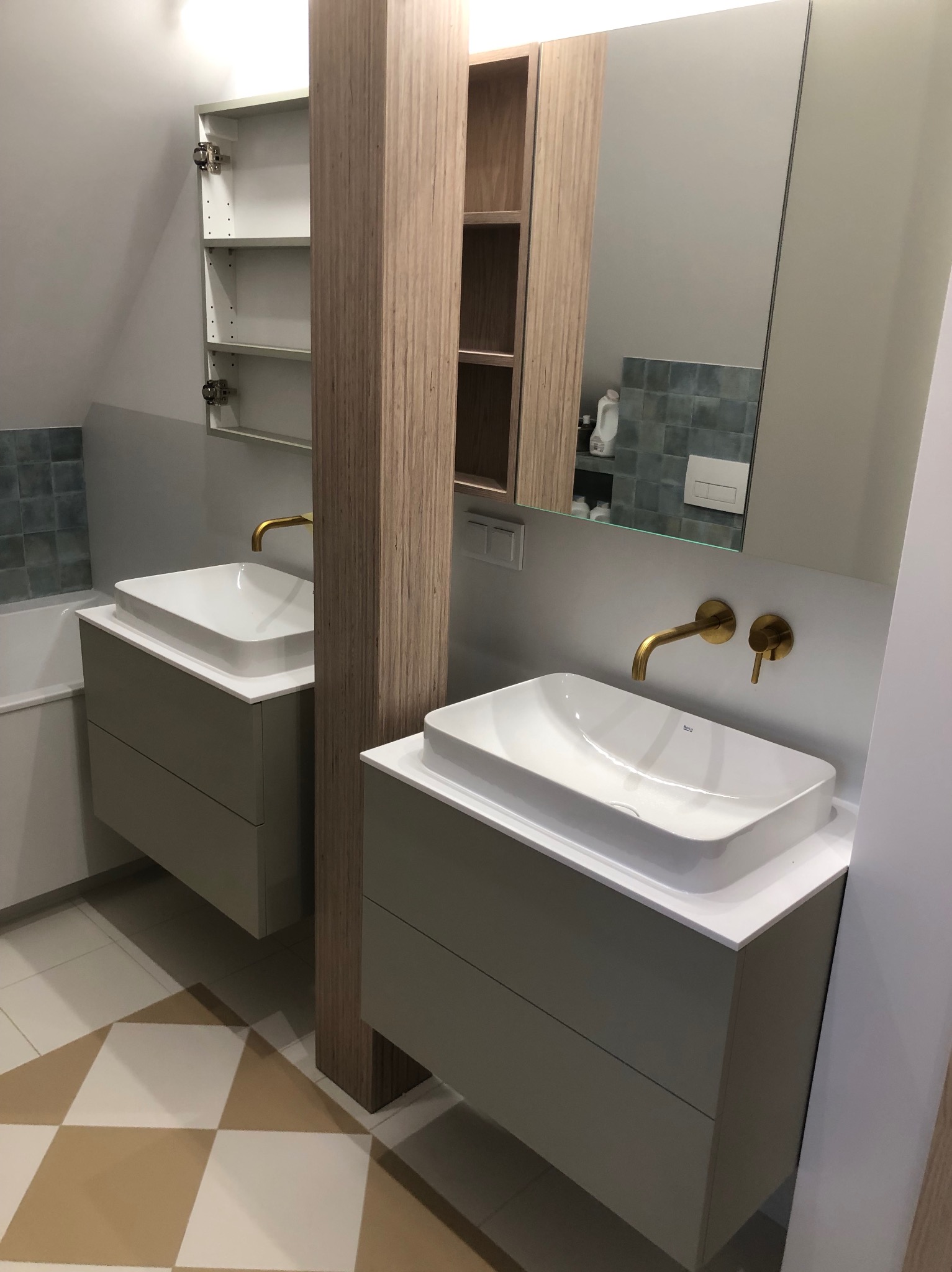 Nowoczesna łazienka z dwoma umywalkami nablatowymi, złotą armaturą i szafkami w odcieniach szarości. Na podłodze wzór w szachownicę. Widoczne lustro i półki.