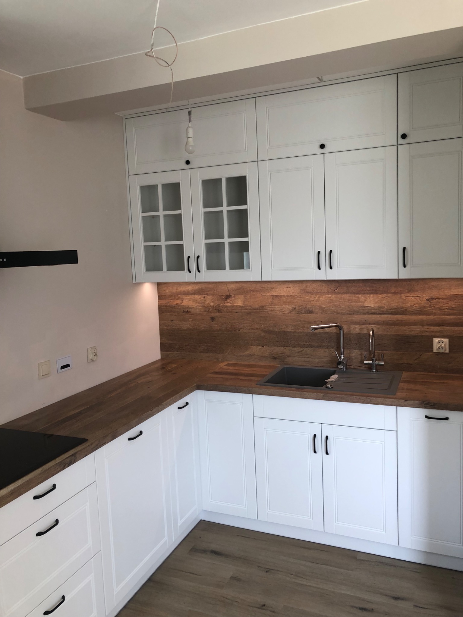 Nowoczesna kuchnia z białymi szafkami z frezowanymi frontami i drewnianym blatem, czarny zlew i bateria, widoczne gniazdka elektryczne na ścianie.