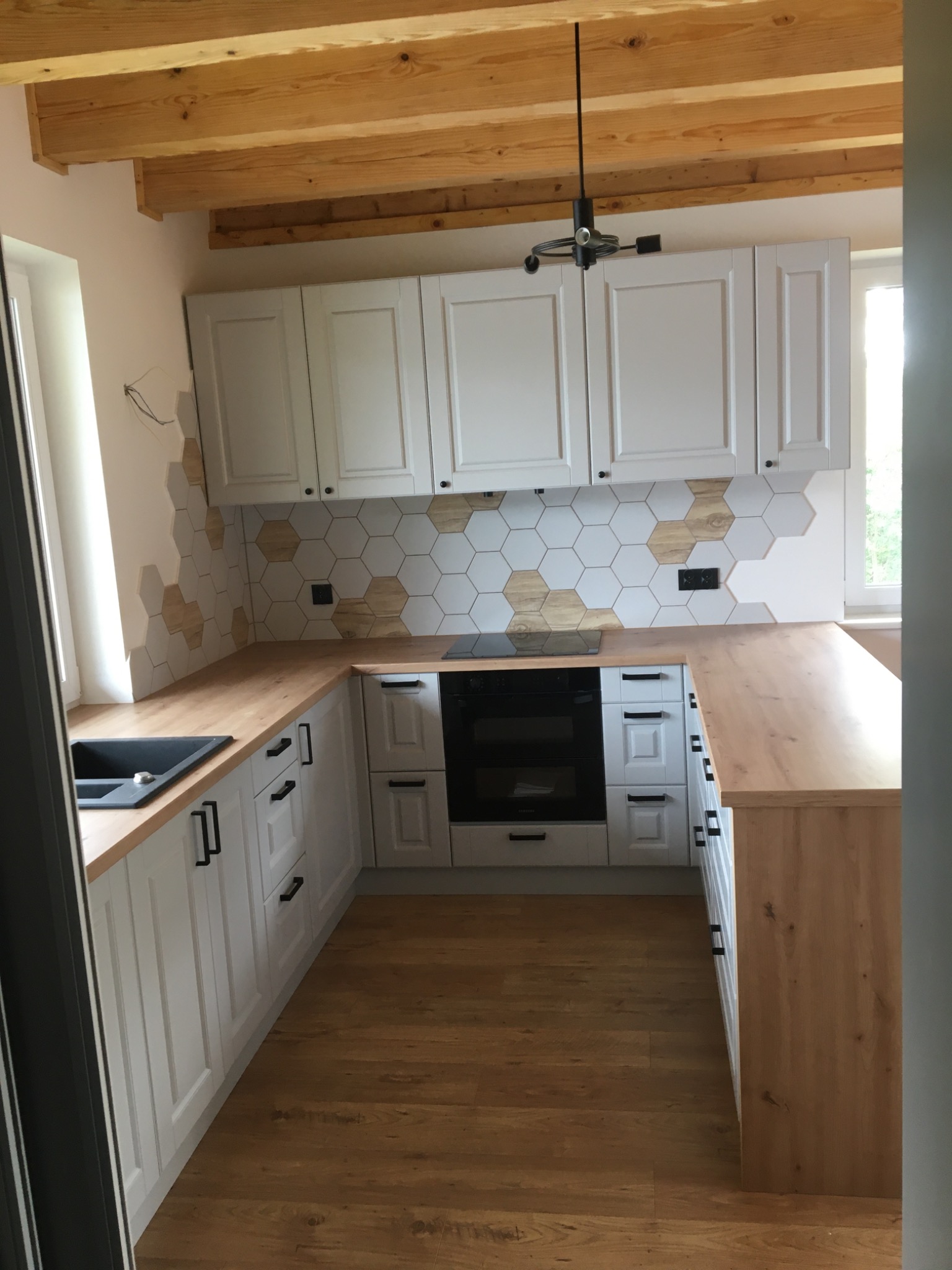 Jasna kuchnia z białymi szafkami, drewnianymi blatami i heksagonalnymi płytkami na ścianie, sufit z drewnianych belek, czarne uchwyty szafek.