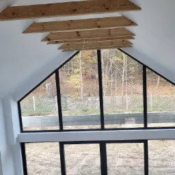 Wnętrze poddasza z drewnianą konstrukcją stropu i dużym oknem z widokiem na jesienny krajobraz, surowy beton na podłodze.