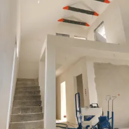 Wnętrze w trakcie prac wykończeniowych: surowe schody betonowe, ściany częściowo pokryte gładzią, belki stropowe zabezpieczone folią, agregat malarski Texspray T-Max 506 na podłodze.