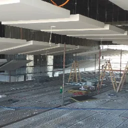 Montaż podwieszanego sufitu z płyt w dużej przestrzeni, widoczne rusztowania i materiały budowlane na metalowej podłodze.