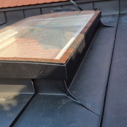 CuDACH Marcin Cuderski - Szczegółowe ujęcie okna dachowego z drewnianą ramą na pokryciu z blachy na rąbek stojący, widoczne dachówki ceramiczne w tle.