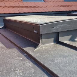 CuDACH Marcin Cuderski - Szczegółowe ujęcie okna dachowego Velux z obróbką blacharską na dachu pokrytym blachą na rąbek stojący, widoczny fragment dachu z dachówką ceramiczną w tle.