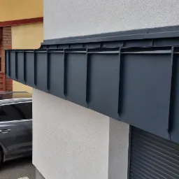 Szare, metalowe orynnowanie dachu nowoczesnego budynku, widoczne z boku, z fragmentem zaparkowanego samochodu w tle.