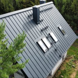 CuDACH Marcin Cuderski - Szary dach z blachy na domu jednorodzinnym, widok z góry, z trzema oknami dachowymi i kominem. Obok drzewo iglaste i częściowo widoczne materiały budowlane.