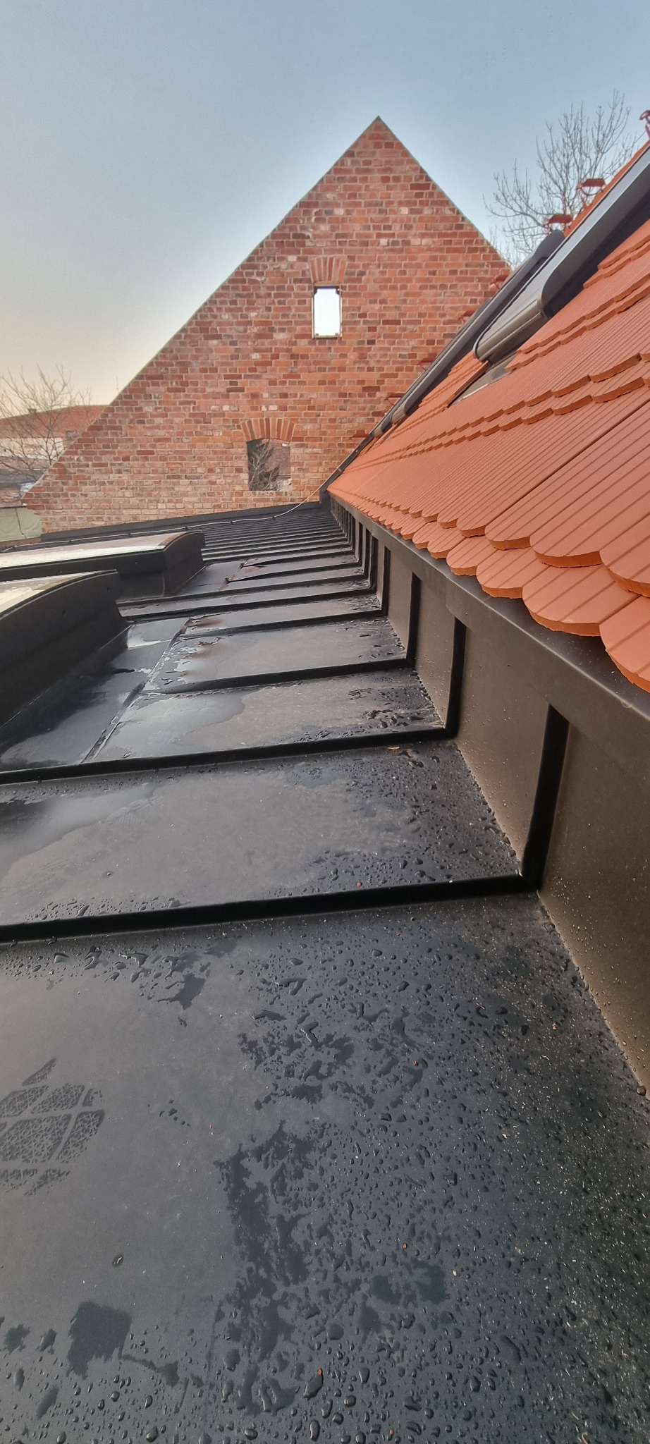 Czarne, mokre panele dachowe z widocznym odciskiem buta, w tle ceglana ściana szczytowa z oknem i fragment dachu z dachówki ceramicznej.