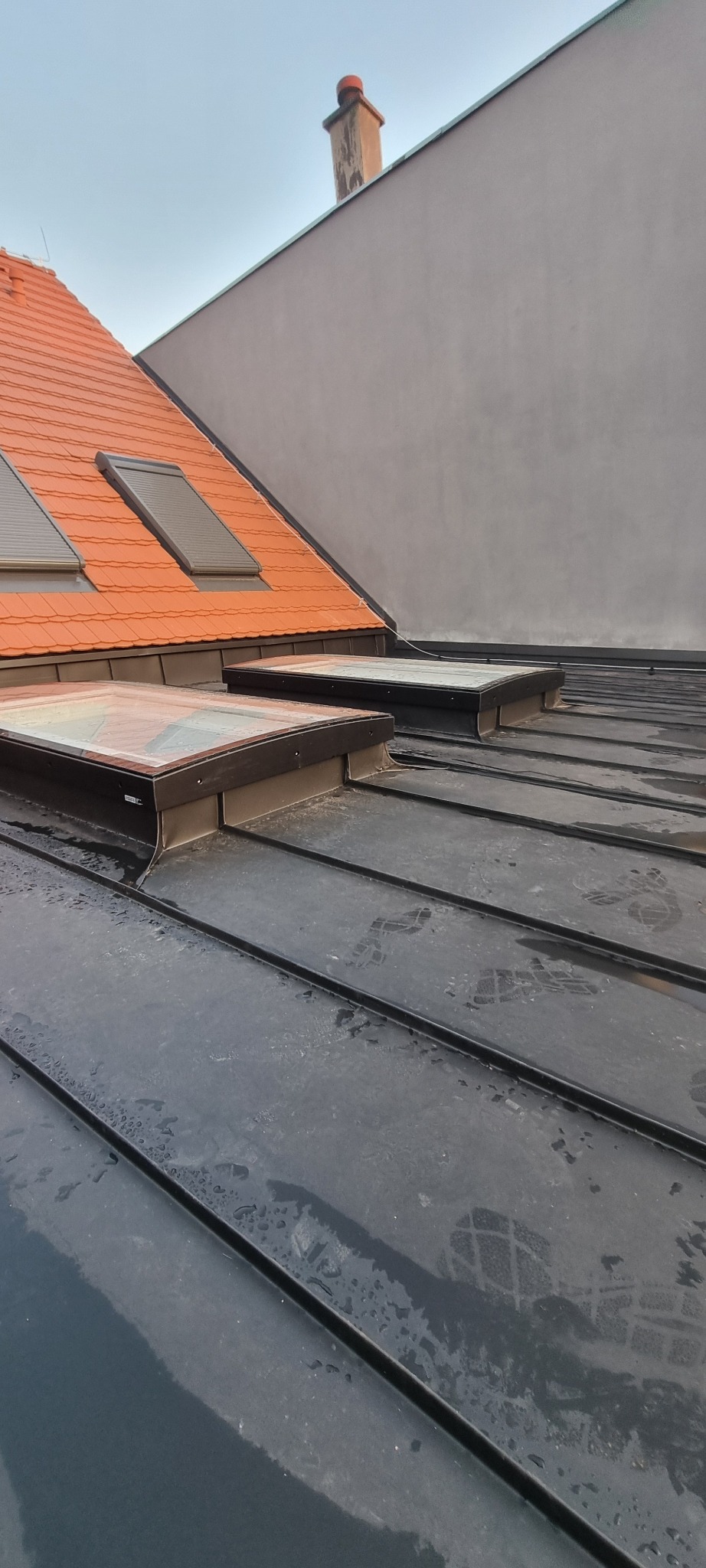 Czarny dach z blachy na rąbek stojący, widoczne okna dachowe i ślady stóp, w tle dachówka ceramiczna i komin