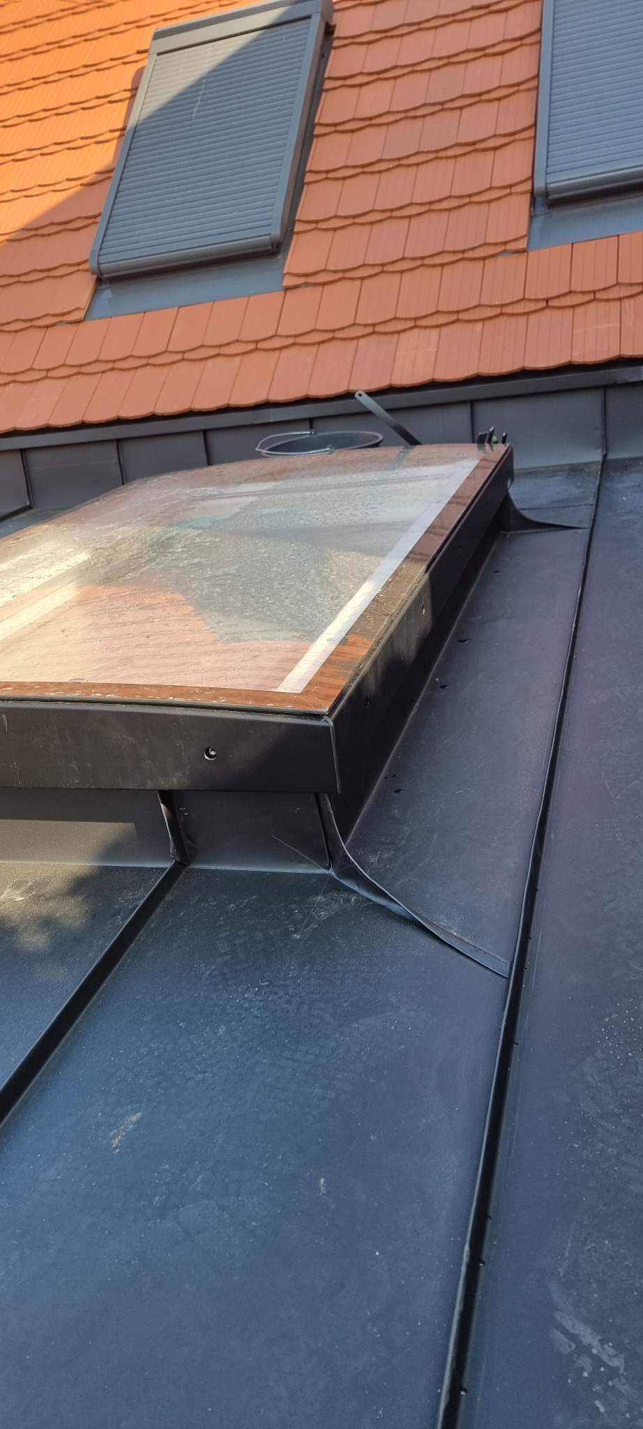 Szczegółowe ujęcie okna dachowego z drewnianą ramą na pokryciu z blachy na rąbek stojący, widoczne dachówki ceramiczne w tle.