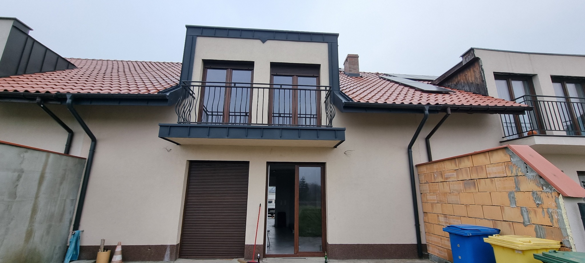 Dom jednorodzinny z czerwonym dachem, ciemnymi obróbkami blacharskimi i balkonem z metalową balustradą, częściowo w trakcie budowy.