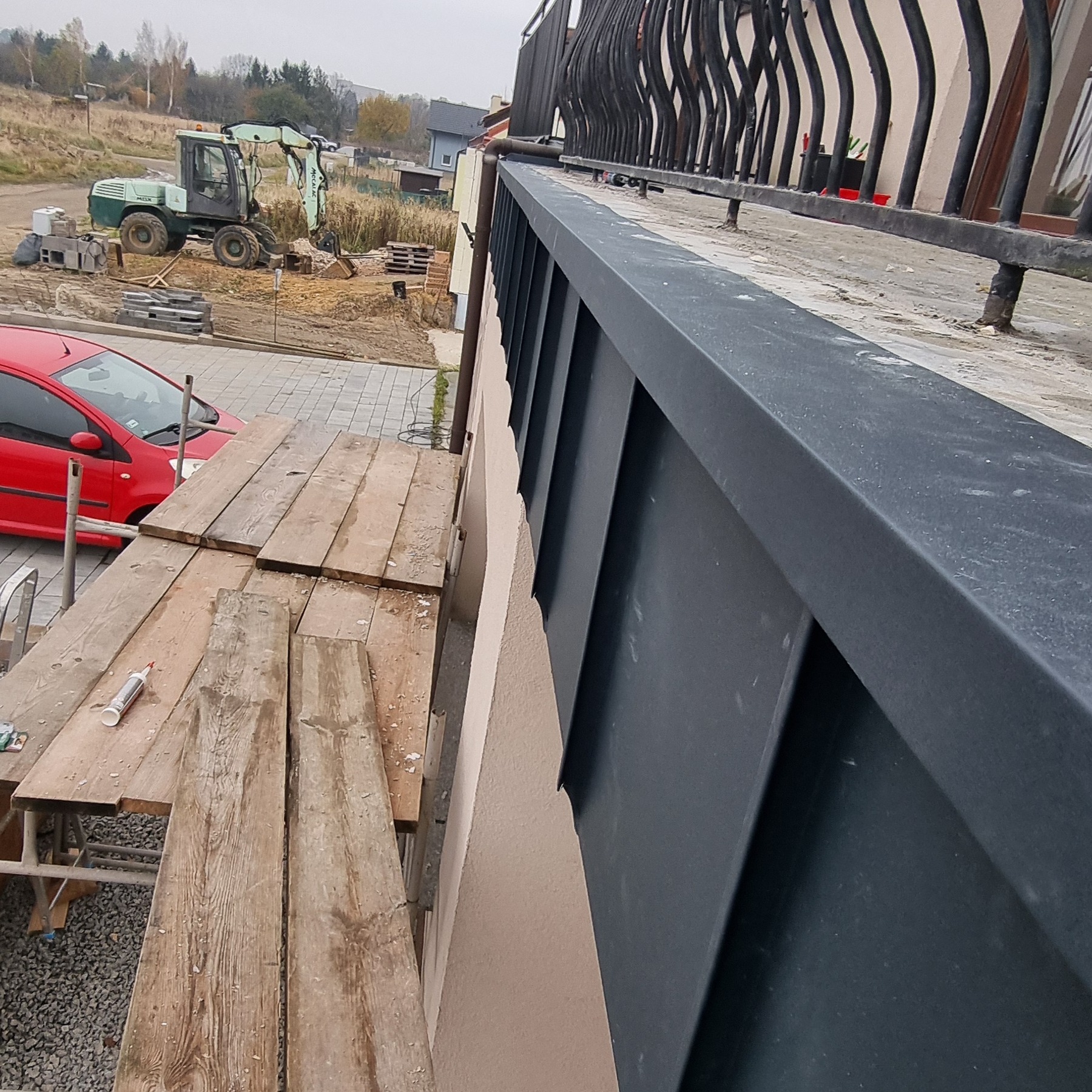 Montaż czarnej metalowej listwy przypodachowej na jasnej elewacji budynku z widocznym rusztowaniem i czerwonym samochodem w tle.