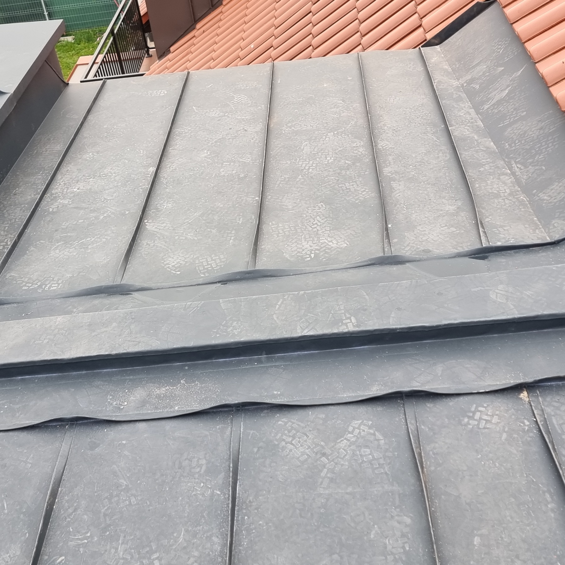 Szare panele blaszane na dachu, widoczne ślady po butach, obok dach z czerwonej dachówki.