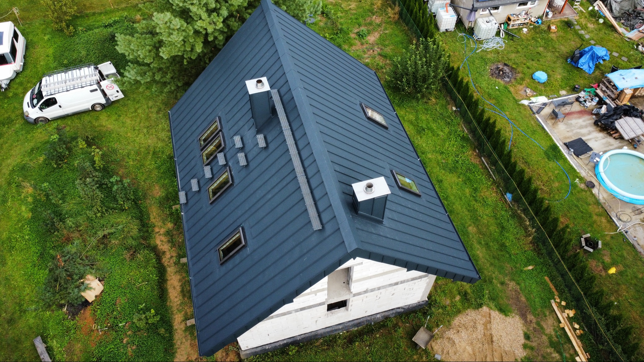 Widok z góry na dom w budowie z ciemnym dachem z blachy na rąbek stojący, z oknami dachowymi i kominami. Obok zaparkowany biały bus z drabiną na dachu.