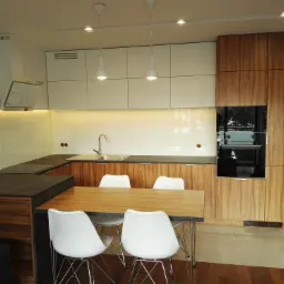 Nowoczesna kuchnia z białymi frontami szafek górnych i drewnopodobnymi frontami dolnych, zintegrowana z jadalnią, oświetlona lampami wiszącymi w kształcie kropli.