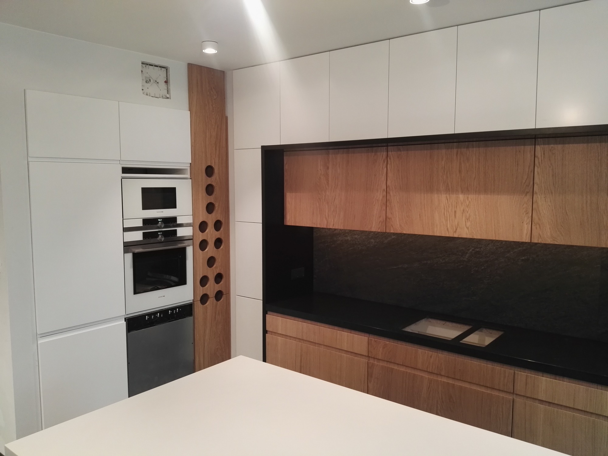 Nowoczesna kuchnia z frontami z jasnego drewna i białymi szafkami, czarny blat z wbudowanym zlewem, drewniany panel z okrągłymi otworami na butelki.