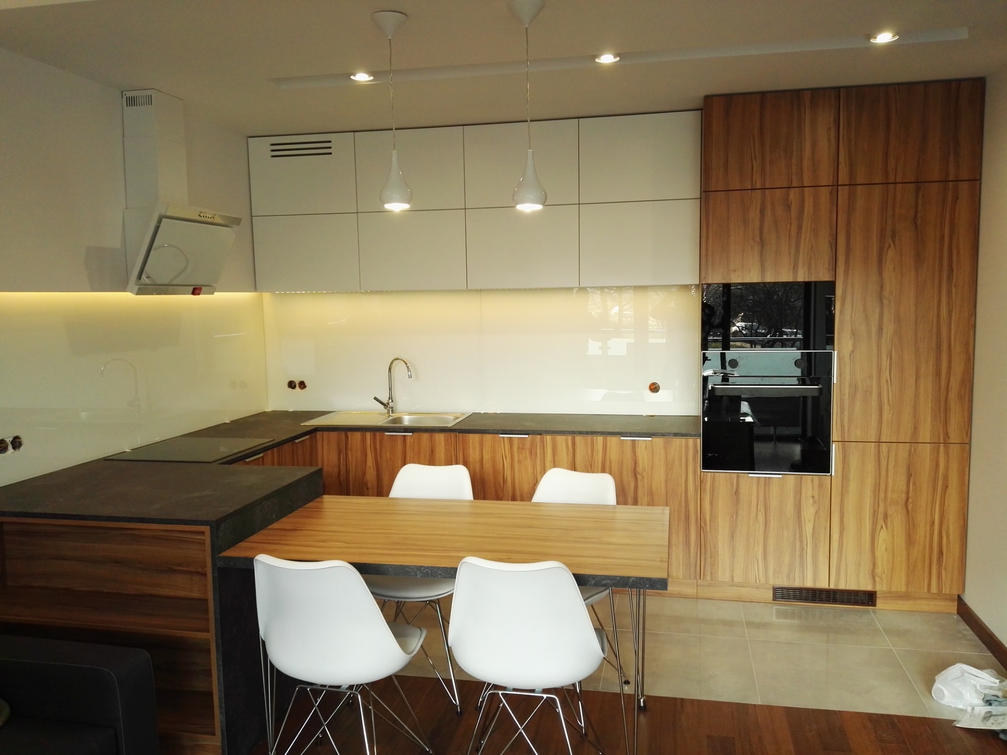 Nowoczesna kuchnia z białymi frontami szafek górnych i drewnopodobnymi frontami dolnych, zintegrowana z jadalnią, oświetlona lampami wiszącymi w kształcie kropli.