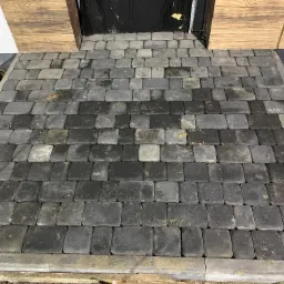 Ułożona szara kostka brukowa przed wejściem do domu, z widocznymi elementami wykończenia drewnianego i szarym cokołem.