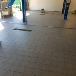 Szara, kwadratowa podłoga z płytek w warsztacie samochodowym z podnośnikiem samochodowym, widoczne ślady użytkowania.