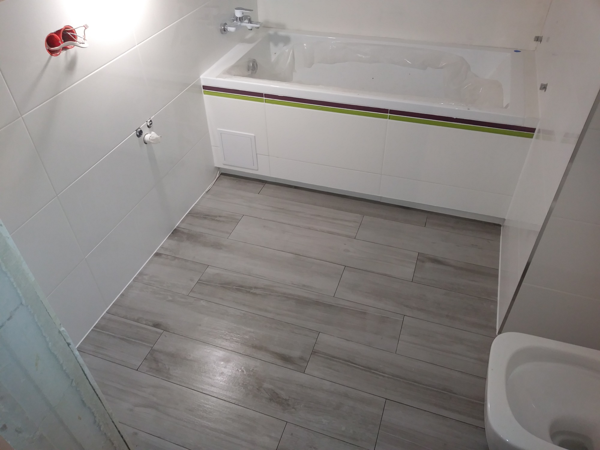 Biała Podlaska: Nowoczesna łazienka po remoncie, z wanną obudowaną białymi płytkami z dekoracyjnym paskiem, szare płytki podłogowe imitujące drewno, białe płytki ścienne.