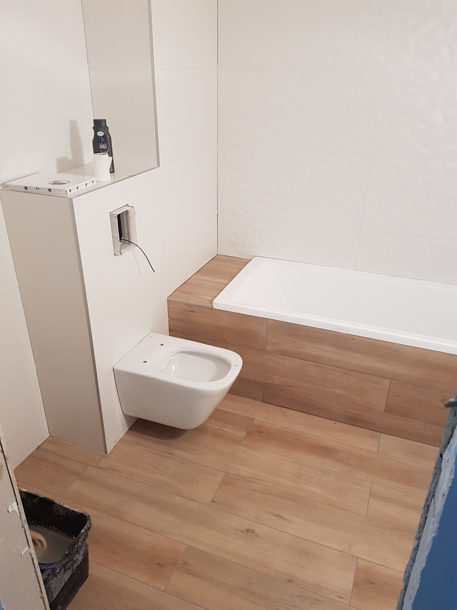 Nowoczesna łazienka w trakcie wykończenia: podwieszana miska WC z instalacją, białe płytki ścienne z delikatną fakturą, obudowa wanny z drewnopodobnych płytek, podłoga wyłożona płytkami imitującymi...