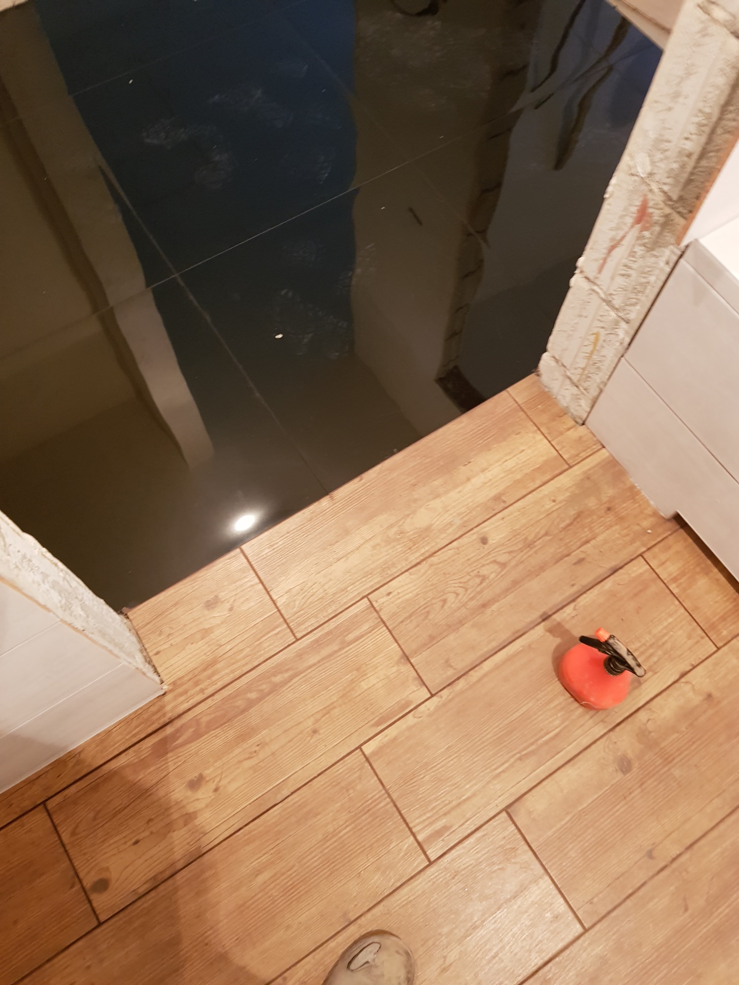 Podłoga w łazience z brązowych płytek imitujących drewno, kontrastująca z nowo położonymi, błyszczącymi, czarnymi płytkami podłogowymi w głębi, widoczna przyssawka do płytek i fragment buta.