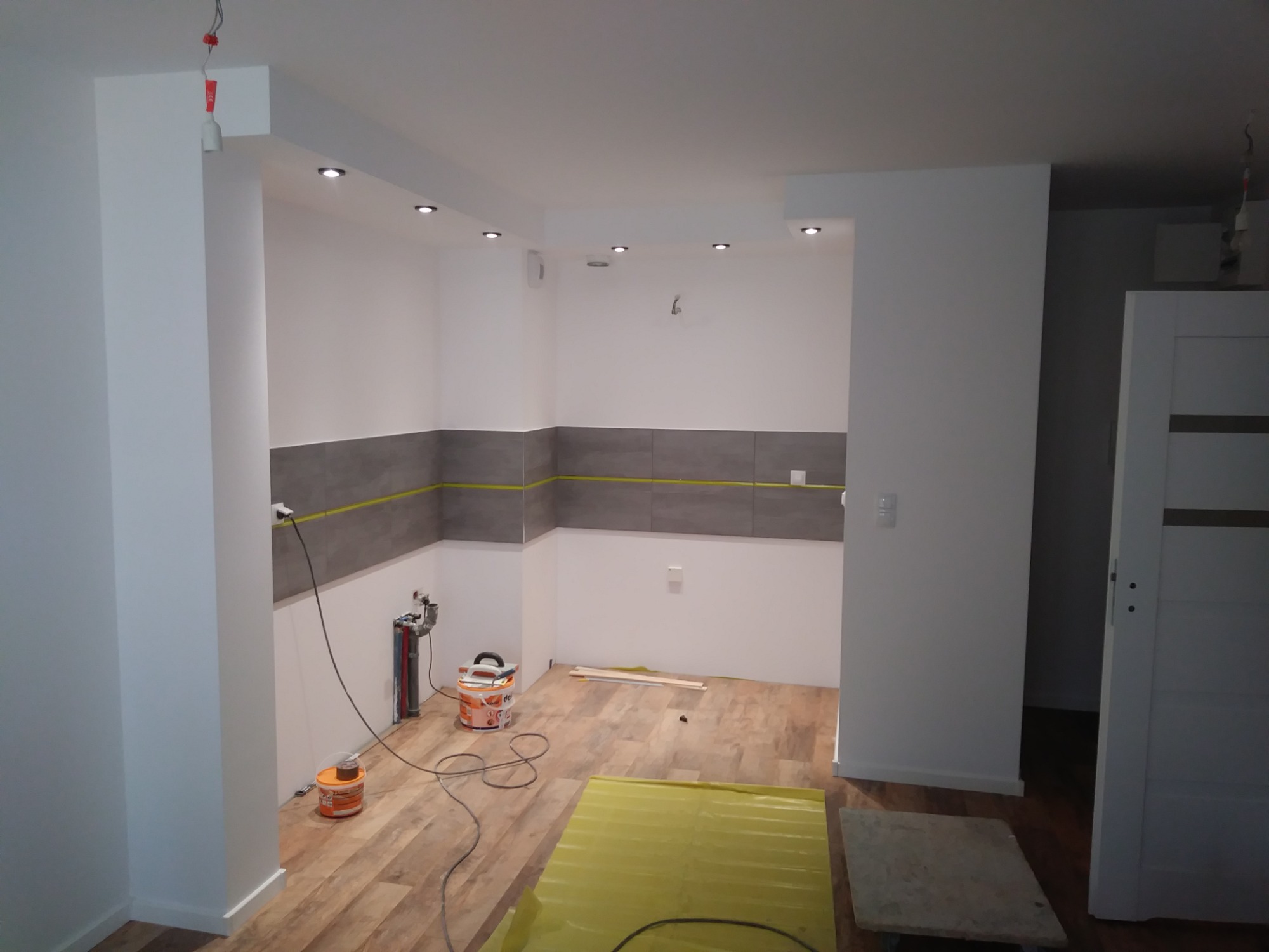 Wnętrze kuchni w trakcie remontu: położone szare płytki, widoczne narzędzia, wiadro z farbą, zabezpieczona podłoga folią, przygotowane listwy przypodłogowe i instalacja wodno-kanalizacyjna.