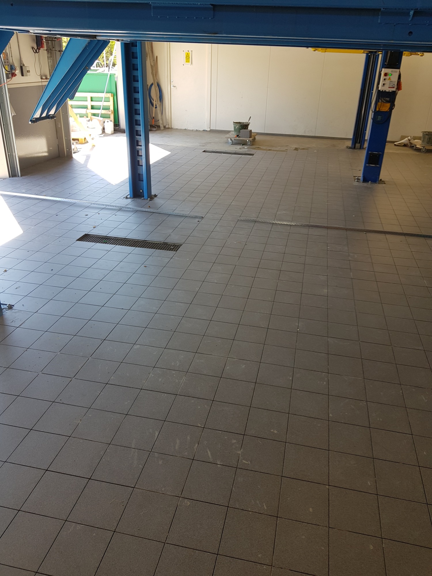 Szara, kwadratowa podłoga z płytek w warsztacie samochodowym z podnośnikiem samochodowym, widoczne ślady użytkowania.