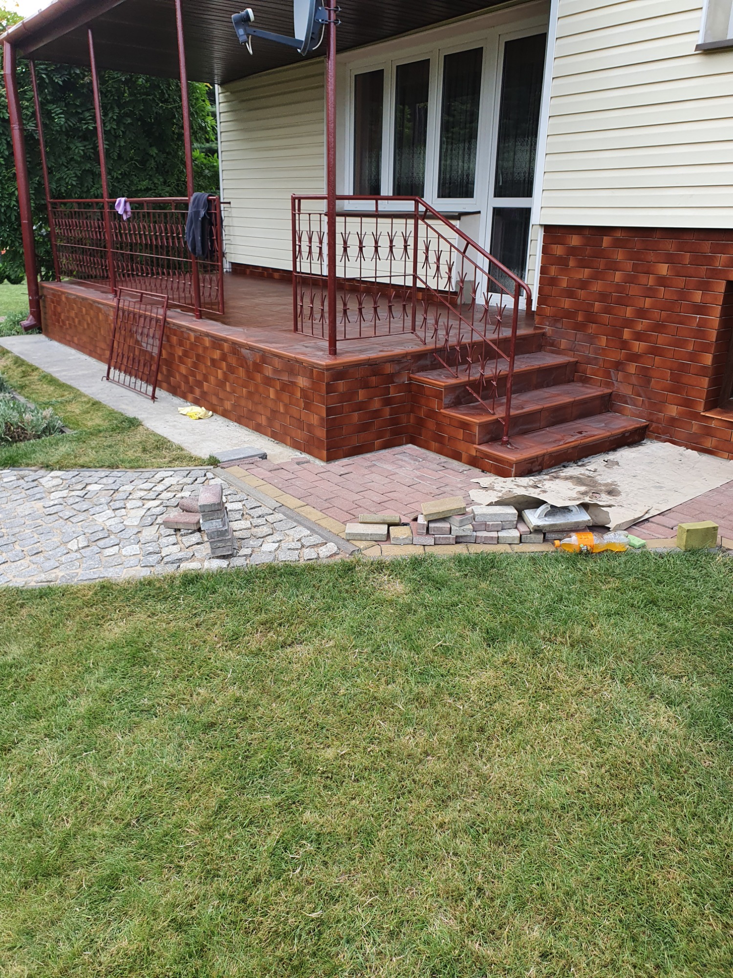 Wykończony taras z ceglaną elewacją i schodami, kontrastujący z niedokończoną ścieżką z kostki brukowej i zielonym trawnikiem.